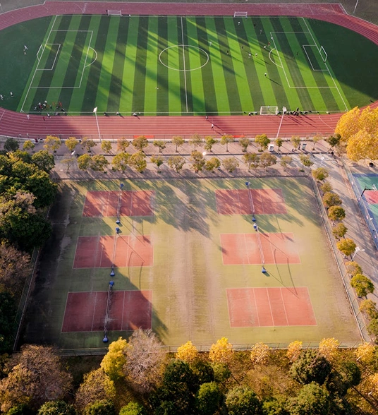 Aerial view of a sports complex featuring a football pitch and several tennis courts, surrounded by trees.