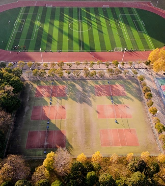 Aerial view of a sports complex featuring a football pitch and several tennis courts, surrounded by trees.