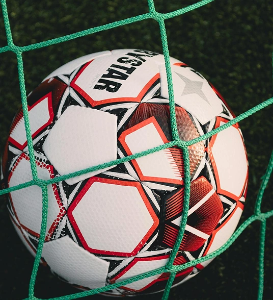 Close-up of a football hitting the net of a goal, with a green field in the background.