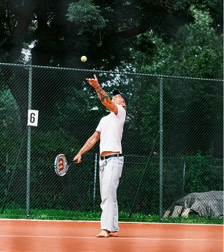 A tennis player serving on an outdoor clay court, surrounded by trees.