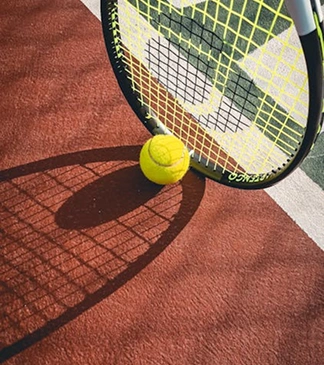 A racket and a tennis ball casting their shadow on the court floor, bathed in soft sunlight.