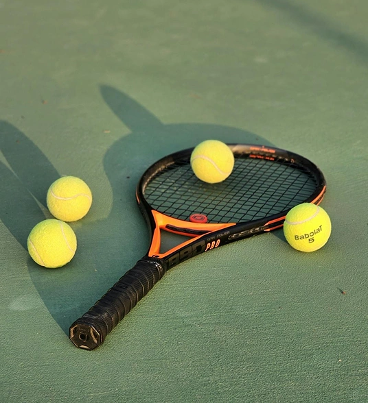 A tennis racket with several tennis balls on the court, under natural sunlight.